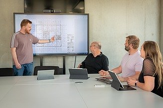 4 Mitarbeiter in einer Besprechung. Ein Mitarbeiter erklärt Plan auf großem Bildschirm. 