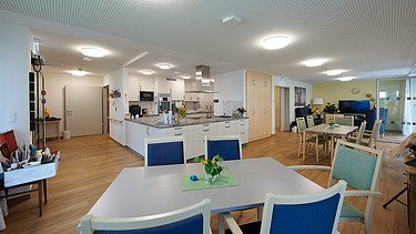 Foto eines Gemeinschaftsraumes mit Tischen und Stühlen und Küche