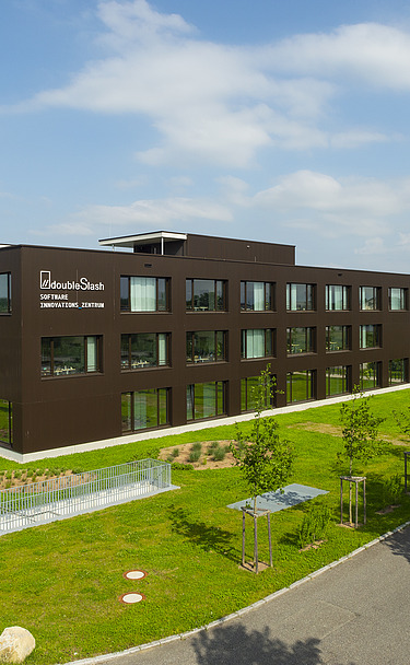 Foto Gebäude von weitem mit brauner Fassade, grüner Wiese und sonnigem Wetter