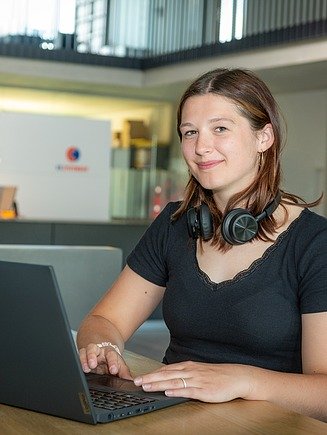 Eine Frau vor einem Stehtisch arbeitet am Laptop. Ein Headset hat sie um den Hals gehängt. 