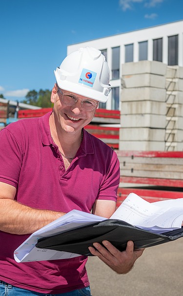 Ein Mann, der einen Bauhelm trägt, schaut sich Baupläne an. Im Hintergrund Schalungen und ein Gebäude