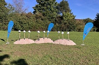 Foto von Spaten mit Helmen auf Erdhügel auf grüner Wiese und Firmenflaggen