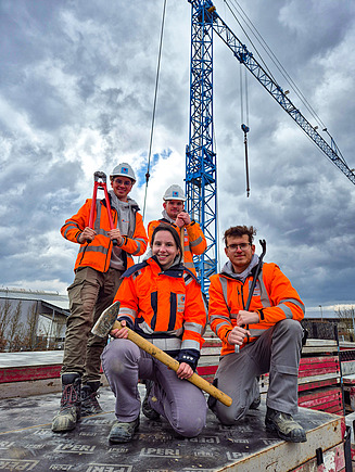 Vier Auszubildende stehen oder knien mit verschiedenen Werkzeugen in der Hand auf Schalungselementen. Im Hintergrund steht ein Kran