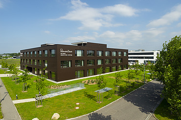 Foto Gebäude von weitem mit brauner Fassade, grüner Wiese und sonnigem Wetter