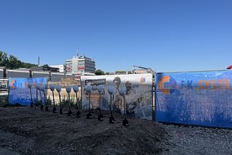 Baufeld mit Bauzaunbanner sowie Spaten und Bauhelme