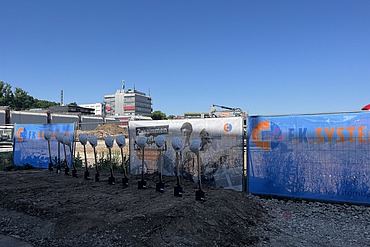 Baufeld mit Bauzaunbanner sowie Spaten und Bauhelme
