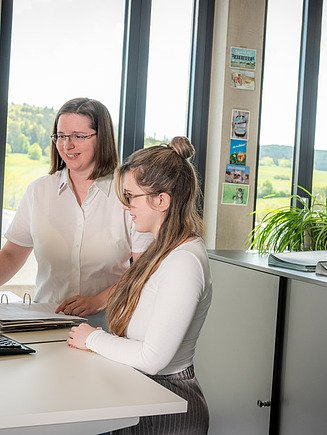 2 weibliche Mitarbeiter stehen am Schreibtisch und schauen zum Bildschirm. Auf dem Schreibtisch liegt ein Ordner. 