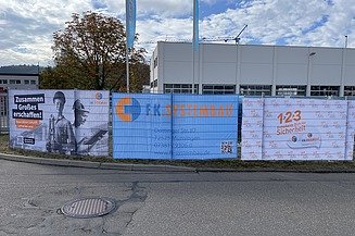 Zaun mit Bannern, im Hintergrund Werkstattgebäude